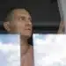 Homem olhando seriamente pela janela com cortina branca ao lado e reflexo de nuvens