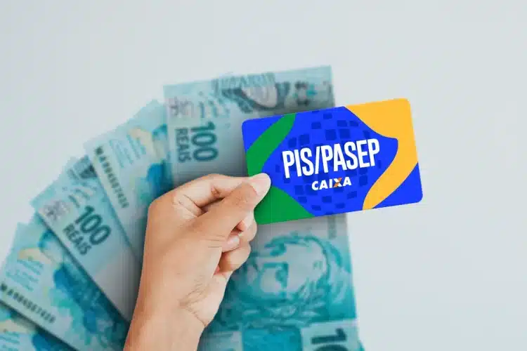 Mão segurando cartão PIS/Pasep da Caixa sobre várias notas de cem reais