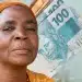 Mulher idosa com lenço laranja posa ao lado de várias notas de cem reais.