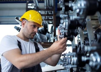 Operário usando capacete amarelo ajusta engrenagens em equipamento industrial.