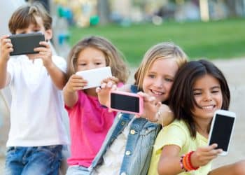 Quatro crianças tirando selfies com smartphones em parque, sorrindo e próximas umas das outras.