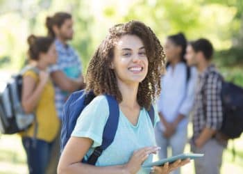 Estudante sorrindo enquanto usa um tablet, com outros colegas ao fundo, representando jovens em busca de oportunidades acadêmicas e profissionais.
