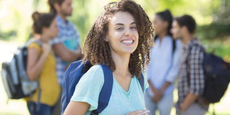 Estudante sorrindo enquanto usa um tablet, com outros colegas ao fundo, representando jovens em busca de oportunidades acadêmicas e profissionais.