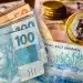 Cédulas de 100 e 50 reais sobrepostas com moedas de 1 real ao fundo.