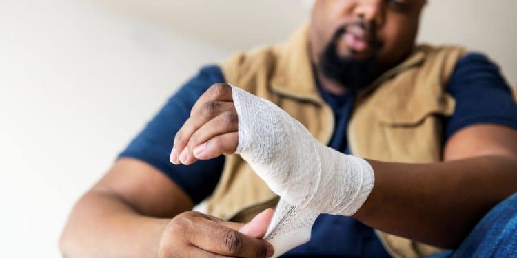 Homem ajustando bandagem na mão ferida com expressão de dor.