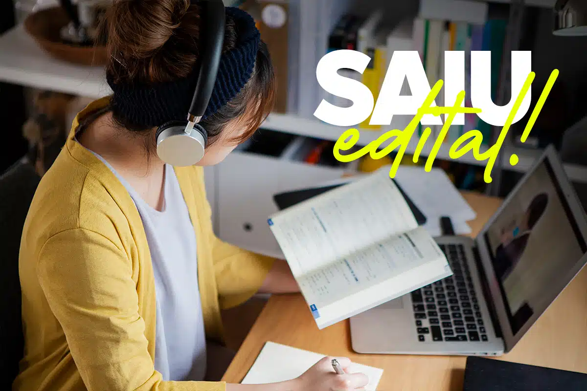Mulher com fone de ouvido estuda com livro aberto e anotações ao lado de notebook.