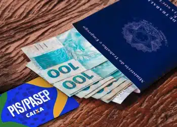 Carteira de trabalho azul, notas de cem reais e cartão PIS/Pasep sobre mesa de madeira