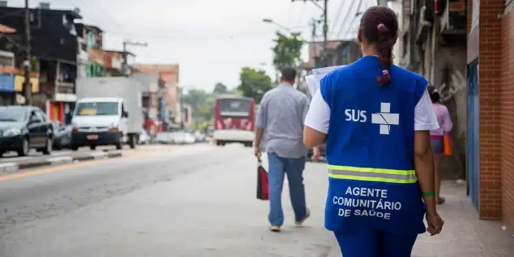 Profissional do SUS caminha em rua de bairro enquanto segura papéis, usando colete azul