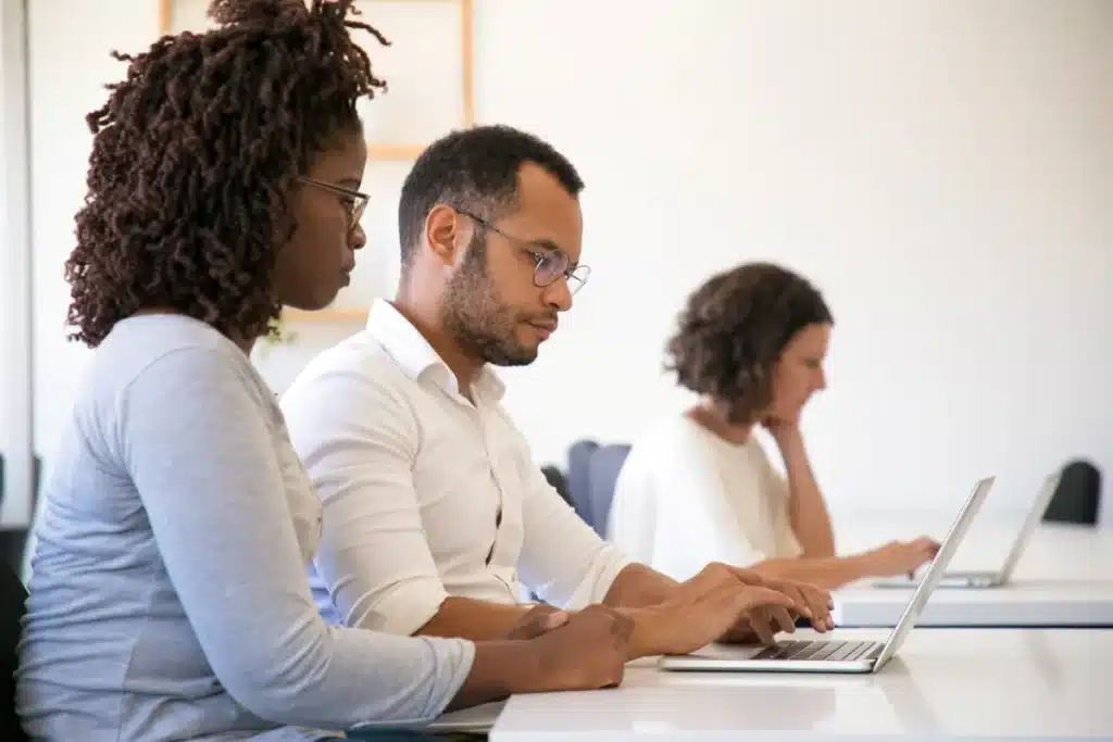 Dois colegas de trabalho interagindo e usando um computador juntos em um ambiente profissional. Um deles está orientando o outro enquanto ambos estão focados nas tarefas.