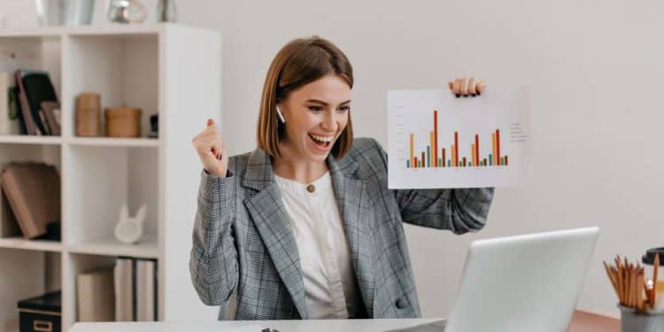 Mulher de negócios comemorando bons resultados de produtividade, segurando gráfico durante videoconferência.