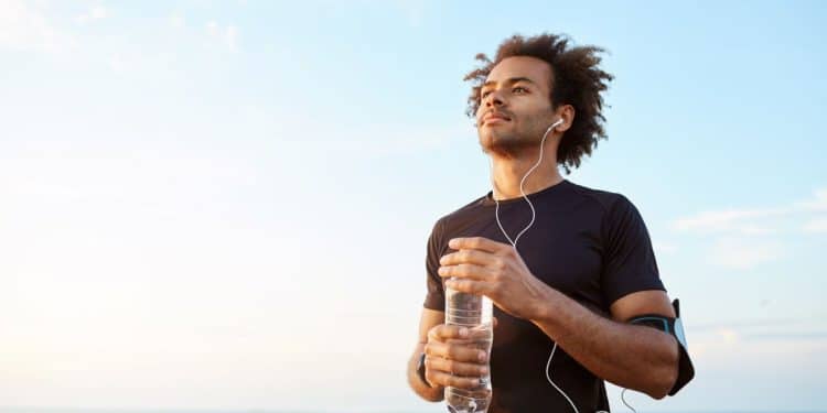 Close de um homem jovem e atlético, de pele negra e cabelo afro, vestindo roupa preta de corrida, com fones de ouvido e uma garrafa de água na mão, olhando pensativo para o horizonte ao pôr do sol em um calçadão à beira-mar.