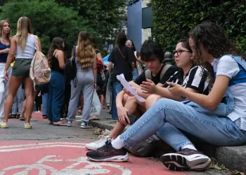 Grupo de estudantes sentados na calçada consultando papéis e celular enquanto outros aguardam em fila na entrada de uma instituição de ensino.