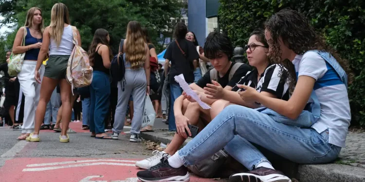 Grupo de estudantes sentados na calçada consultando papéis e celular enquanto outros aguardam em fila na entrada de uma instituição de ensino.