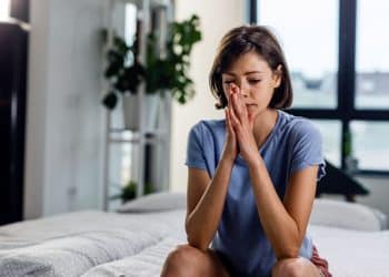 Mulher sentada na cama com as mãos juntas em sinal de preocupação, representando reflexão sobre carência emocional.