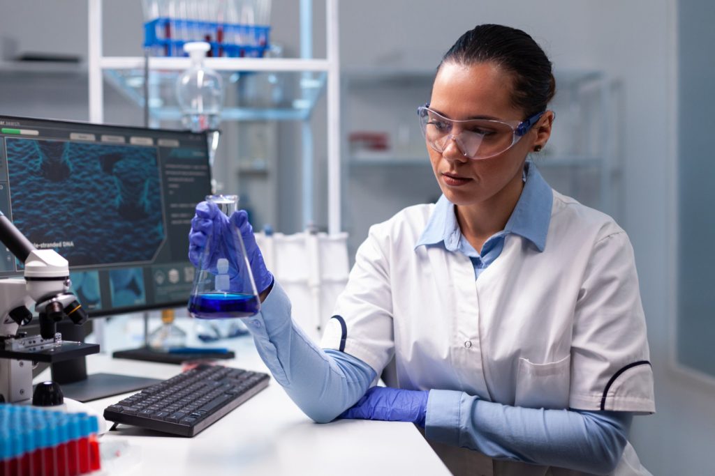 Pesquisadora em laboratório com jaleco branco e óculos de proteção analisando amostra — carreira e salário de R$ 9 mil no concurso IPA SP 2026