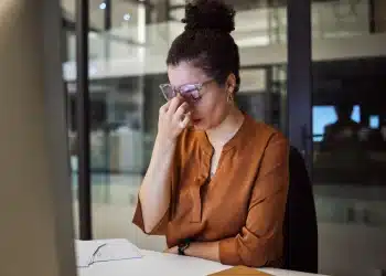Mulher com dor de cabeça, claramente sobrecarregada de trabalho, em escritório com documentos em cima da mesa, expressão de frustração e cansaço.
