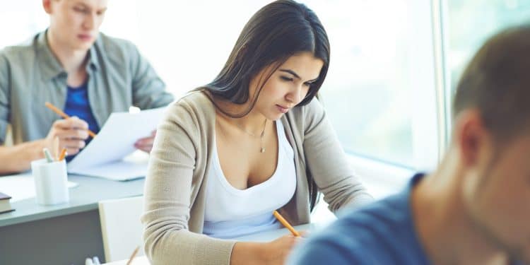 Jovem estudante de cabelo escuro concentrada escrevendo com lápis em caderno em sala de aula com outros estudantes ao fundo