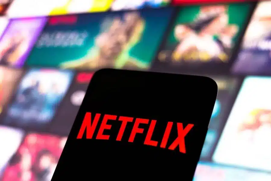Um smartphone com a tela escura exibe o logotipo vermelho da Netflix em destaque, contra um fundo desfocado de outras capas de filmes e séries.