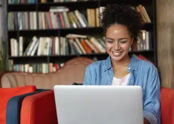 Estudante sorrindo ao usar notebook em biblioteca, acessando portal de certificados