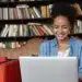 Estudante sorrindo ao usar notebook em biblioteca, acessando portal de certificados