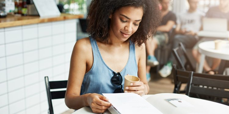 Jovem sorrindo enquanto lê material de estudo em cafeteria com livro aberto e bebida na mão representando preparação para concurso de nível médio