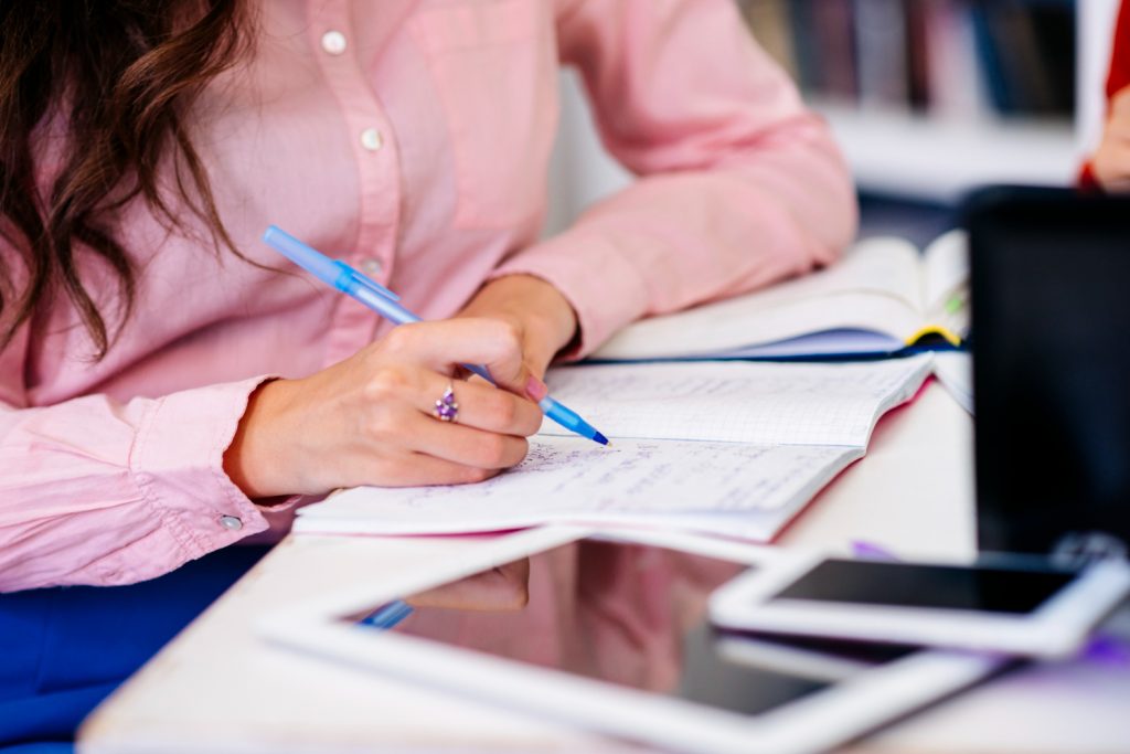 Mulher de camisa rosa anota conteúdos em caderno aberto durante sessão de estudos para concurso público
