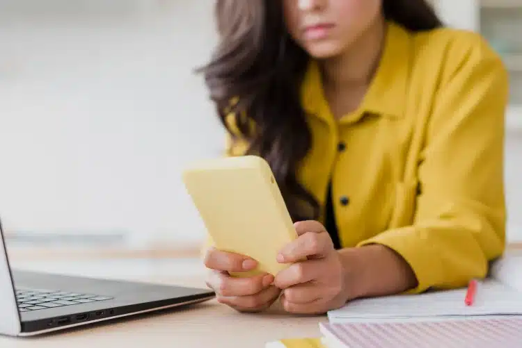 Mulher segura celular amarelo ao lado de notebook e cadernos abertos na mesa
