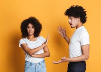 Fotografia de um homem e uma mulher sobre fundo amarelo. A mulher, à esquerda, tem os braços cruzados e uma mão estendida em sinal de "pare", com expressão séria. O homem, à direita, gesticula como se estivesse dando uma explicação ou conselho.