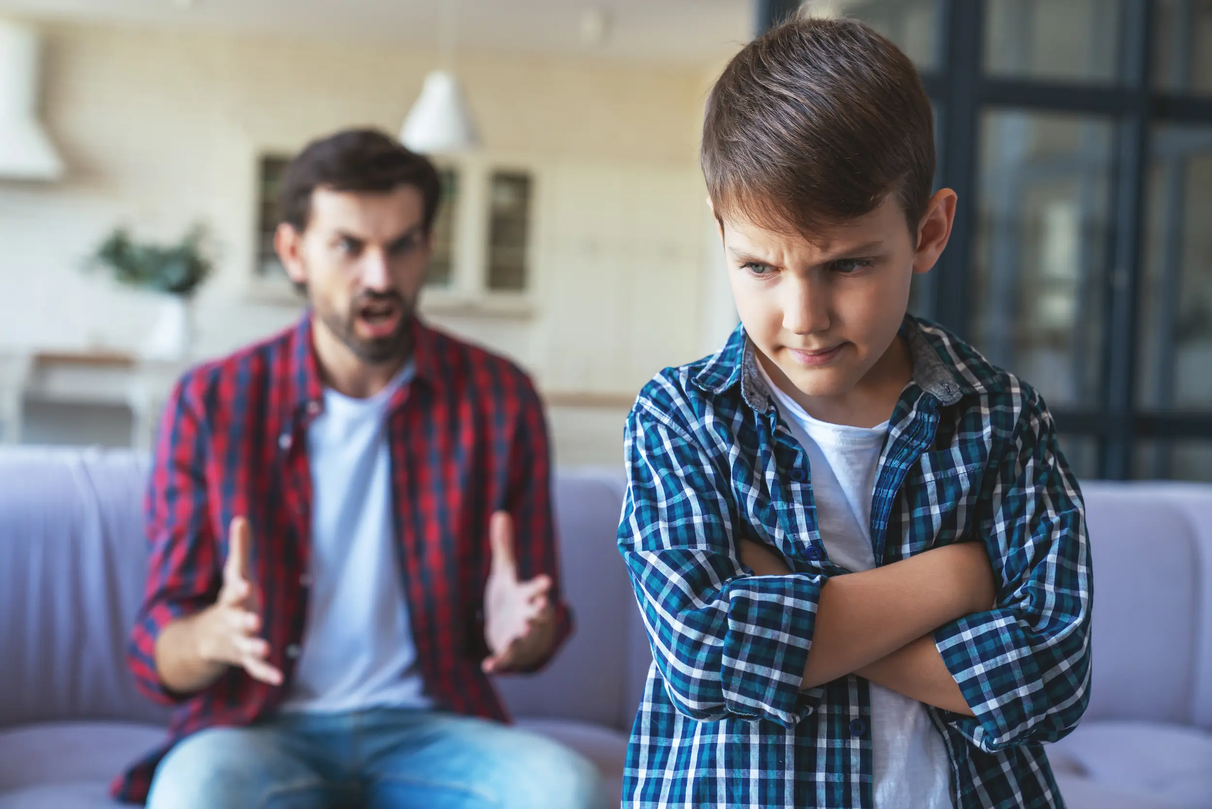 Menino com expressão fechada e braços cruzados enquanto o pai fala de forma dura ao fundo mostrando tensão emocional.