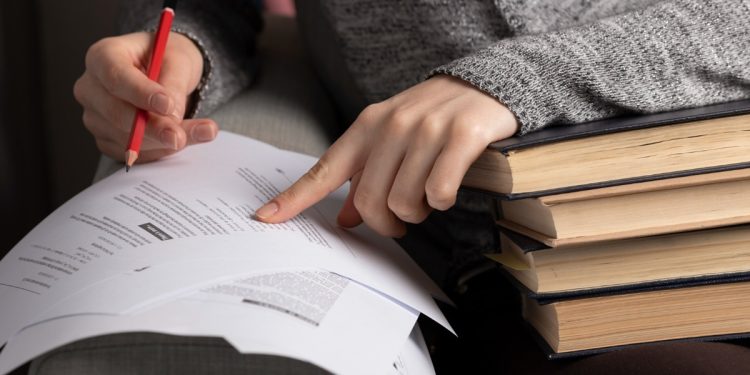 Candidata anotando com lápis vermelho em documentos apoiados sobre pilha de livros durante preparação para o concurso de professor da UFMG com salário de 13,2 mil reais