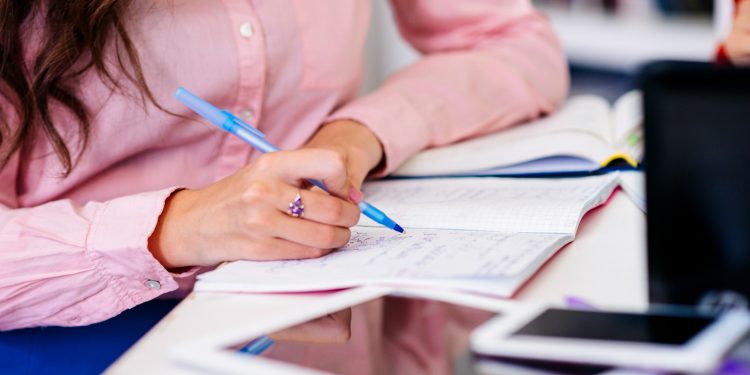 Mão feminina com camisa rosa fazendo anotações em caderno aberto com caneta azul sobre mesa com tablet e smartphone ao lado