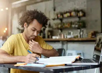 Jovem escreve em caderno enquanto consulta livro aberto ao lado de notebook em mesa