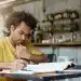 Jovem escreve em caderno enquanto consulta livro aberto ao lado de notebook em mesa