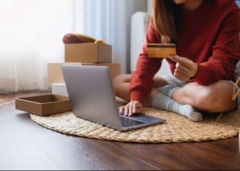 Mulher sentada no chão usando notebook e cartão de crédito rodeada de caixas de encomendas