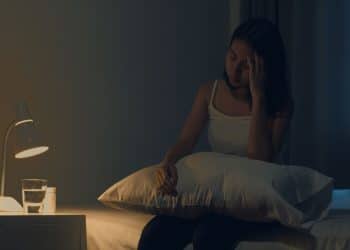 Mulher sentada na cama à noite demonstrando dificuldade para dormir e sinais de cansaço.
