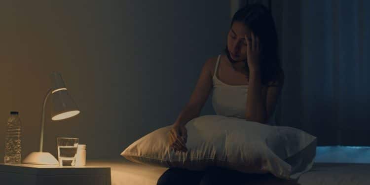 Mulher sentada na cama à noite demonstrando dificuldade para dormir e sinais de cansaço.