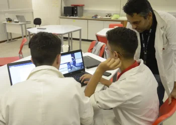 Dois jovens estagiários trabalhando em laptops em uma sala, com um supervisor de jaleco branco orientando, representando o ambiente de estágio profissional e os direitos trabalhistas propostos pelo PL 6350/25.