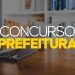 Notebook sobre mesa de madeira com estante ao fundo e texto "Concurso Prefeitura" em destaque, representando o edital do concurso público da Prefeitura de Tremembé SP 2026