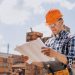Homem com capacete laranja e óculos de proteção analisando uma planta de construção em um canteiro de obras ao ar livre.