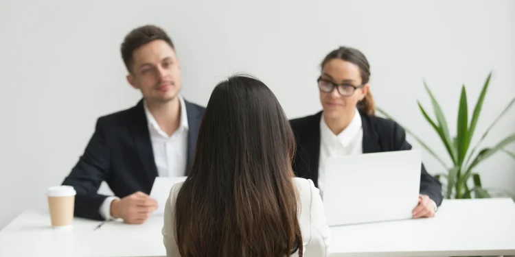 Candidata vista de costas durante entrevista de emprego com dois recrutadores em ambiente corporativo, ilustrando a situação que o simulador do SENAI ajuda a treinar