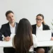 Candidata vista de costas durante entrevista de emprego com dois recrutadores em ambiente corporativo, ilustrando a situação que o simulador do SENAI ajuda a treinar