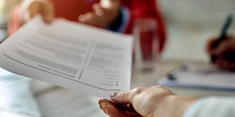 Close nas mãos de um recrutador de terno cinza entregando um currículo impresso a um candidato de regata preta, durante uma entrevista de emprego em um escritório moderno. Ao fundo, uma mesa com um computador e uma planta.