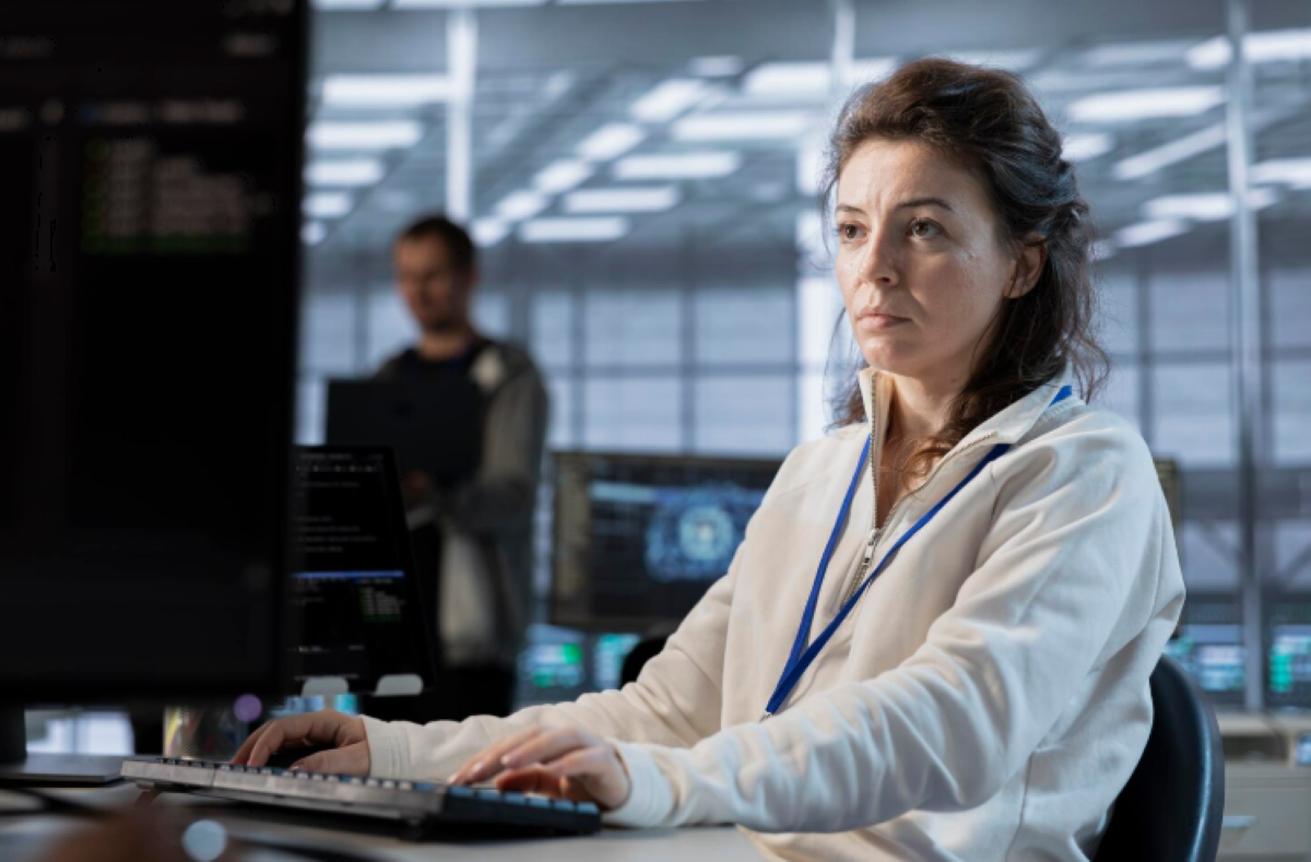 Mulher de casaco branco e crachá azul operando concentrada um teclado em frente a monitores de computador com códigos e interfaces de rede, em um centro de operações de segurança cibernética (SOC), com colega ao fundo.