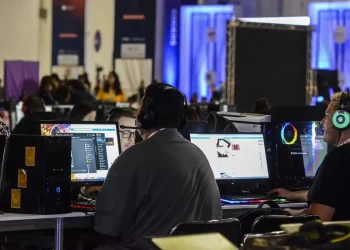 Grupo de profissionais com fones de ouvido trabalhando em computadores desktop durante evento de tecnologia, com iluminação azul ao fundo e diversas estações de trabalho ao longo do ambiente