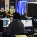 Grupo de profissionais com fones de ouvido trabalhando em computadores desktop durante evento de tecnologia, com iluminação azul ao fundo e diversas estações de trabalho ao longo do ambiente