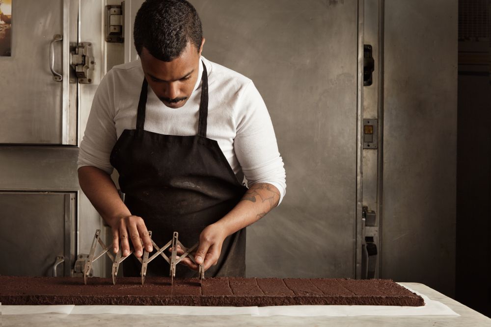 Homem com avental preto e camisa branca trabalhando em uma linha de produção de chocolate, utilizando uma ferramenta de corte extensível para medir porções precisas sobre uma bancada.