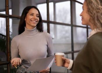 Duas mulheres conversando no trabalho representando críticas veladas e tentativas de desmerecimento de pessoas invejosas.