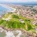 Vista aérea mostra praia extensa, morros verdes e bairros residenciais da cidade litorânea