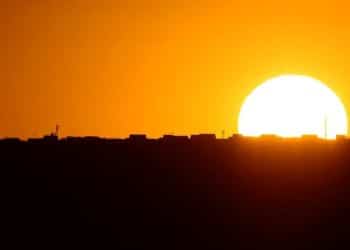 Fotografia de paisagem urbana ao pôr do sol ou nascer do sol. O céu está predominantemente em tons intensos de laranja, amarelo e ouro. À direita, um sol circular muito grande, branco e brilhante, começa a se esconder atrás da linha do horizonte. O horizonte é uma silhueta preta e sólida de edifícios, prédios baixos e antenas de comunicação, sem detalhes visíveis. A parte inferior da imagem é composta por uma área escura, preta e indefinida.