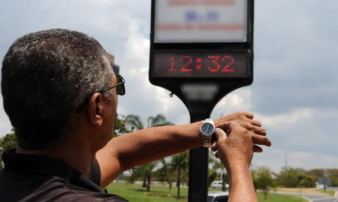 Close-up de um homem de meia-idade, com cabelos grisalhos e óculos de sol, visto de perfil em um dia ensolarado. Ele usa uma camiseta preta. Seus dois braços estão estendidos para a direita da imagem. Com a mão direita, ele segura e ajusta a coroa de um relógio de pulso analógico de prata no pulso esquerdo. Ao fundo, ligeiramente desfocado, um totem de relógio de rua digital mostra a hora '12:32' em LEDs vermelhos brilhantes contra um céu claro com nuvens brancas e palmeiras verdes ao longe.
